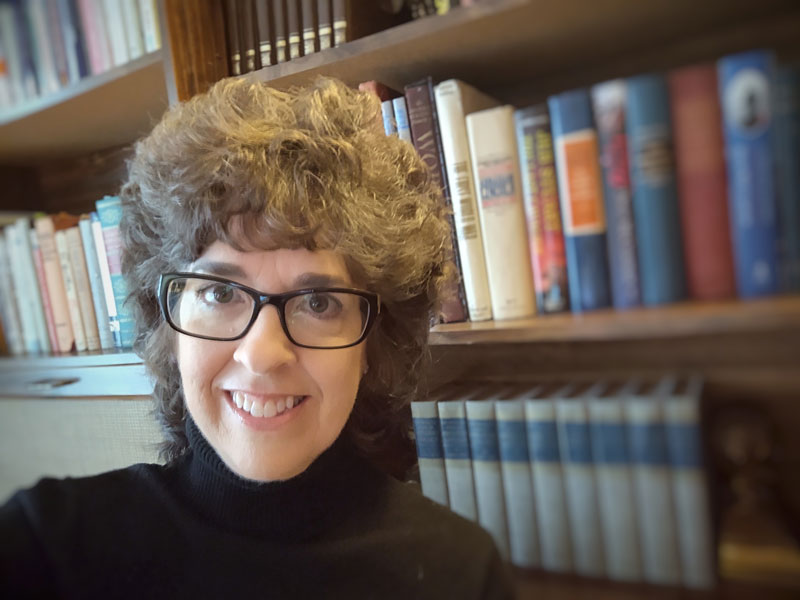 Writer, author, and publisher Melissa Newton smiling in front of her bookcase. She is wearing her signature dark glasses and a black turtle neck.